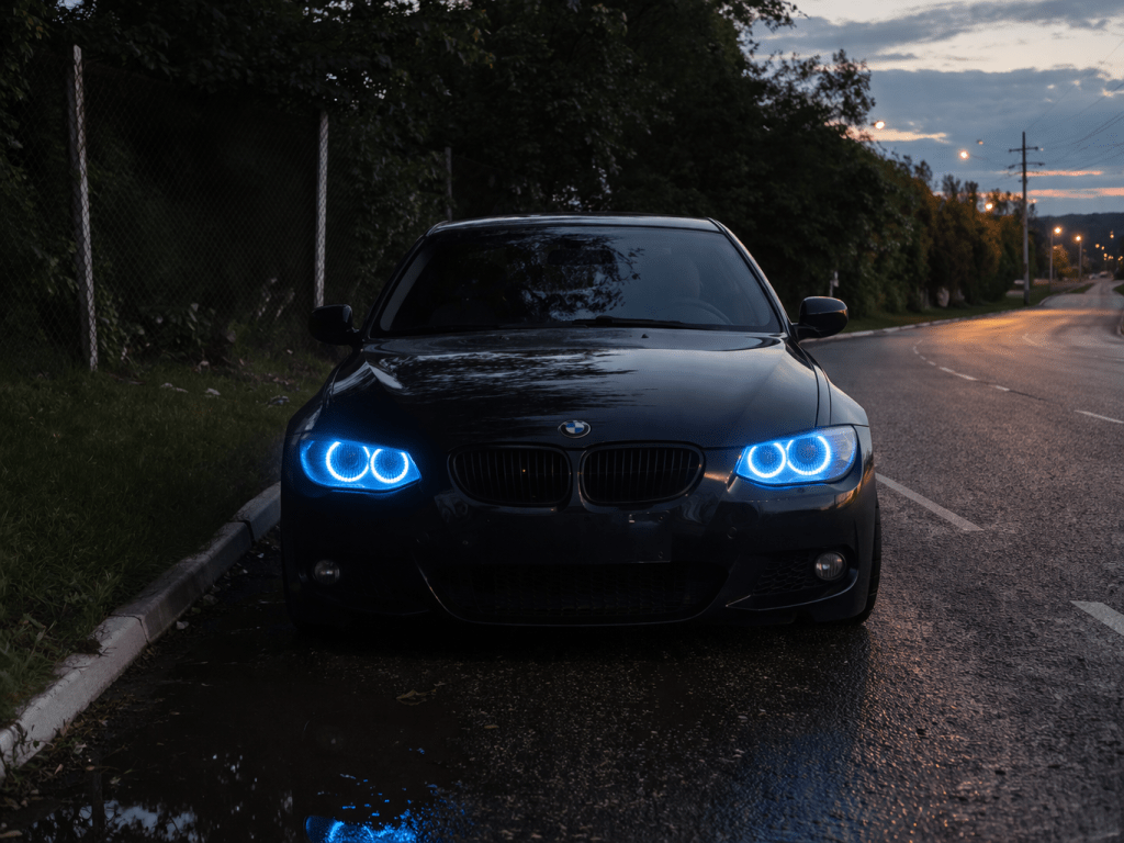 Carro BMW preto com faróis azuis em uma rua à noite, refletindo em poças de água.